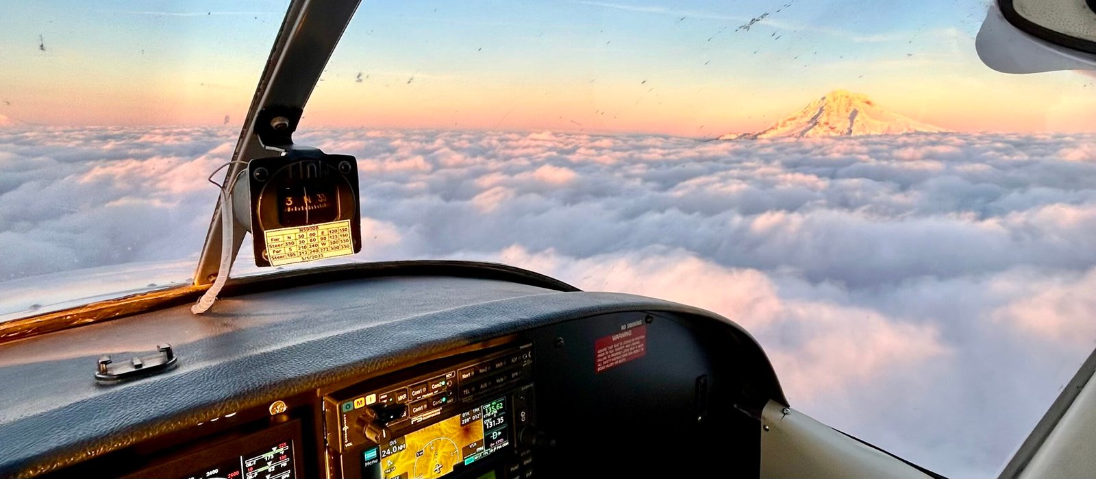 Cockpit view of a small aircraft flying above fluffy clouds, with a snow-capped mountain and a colorful sunset in the background.